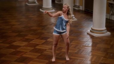 Mavi gecelikli çıplak ayaklı bir dansçının duygusal performansı geniş bir dans salonunun parke zemininde akışkan hareketler gösteriyor.