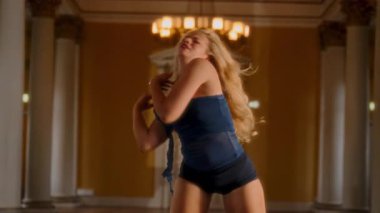 Sarışın bir kadın, büyük bir salonda klasik sütunlarla şiddetli bir şekilde dans ediyor. Güçlü bir enerji ve zarif bir yoğunluk sergiliyor.