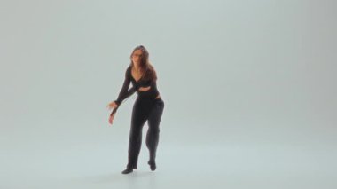 Siyahlı genç bir kız, minimalist beyaz bir stüdyoda dans ediyor, akıcı ve anlamlı hareketler sergiliyor.