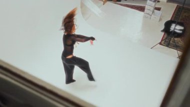 Siyahlı kendine güvenen bir kız minimalist beyaz bir stüdyoda yüksek açılı bir çekimde etkileyici bir ters takla atıyor.