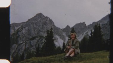 Çimenli bir yamaçta oturan bir kadının görüntüsü. Arkasında heybetli kayalık kayalıklar yükseliyor.