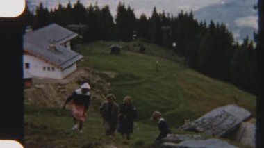 8mm 'lik klasik bir fotoğraf. Büyüleyici bir dağ kulübesinin yanından geçen bir aile. Etrafı çam ağaçlarıyla çevrili.