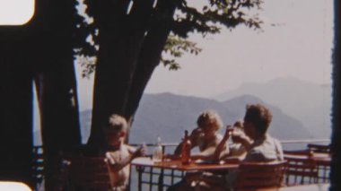 8mm 'lik klasik bir aile görüntüsü arka planda görkemli dağ siluetleriyle birlikte açık bir masanın etrafında toplanmış, birlikte içiyor ve eğleniyorlardı.