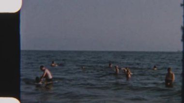 Mutlu insanlar kumsalda güneşli bir günün tadını çıkarıyorlar. Yumuşak dalgalar açık mavi gökyüzünün altında kıyıya vuruyor.