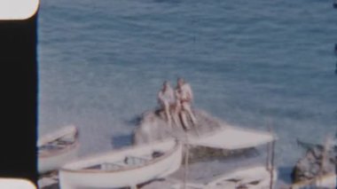 İki kız, deniz kıyısındaki büyük bir kayanın üzerinde huzur içinde oturur. Kayıklar yavaşça kıyıya yanaşır ve sakin sahil manzarasının tadını çıkarırlar.