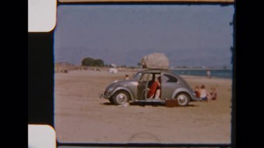 Bir aile, Terracina Sahili 'nde güneşli bir gezinin tadını çıkarıyor. Sahilde park etmiş klasik bir Beetle arabası ve arka planda rahatlatıcı okyanus dalgaları var.
