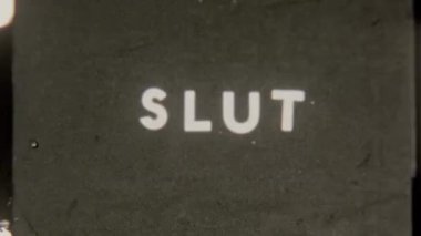 Grainy 8mm film footage displaying the word SLUT in bold all caps on a textured black background, capturing a retro, vintage aesthetic