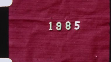 Grainy 8mm film çerçevesi 1985 'in yılını görünür film dokusu ve çıkıntı delikleriyle sıkıntılı bir kırmızı arka planda gösteriyor.