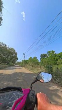 Bereketli yeşillik ve yaprakların arasından süzülen yumuşak güneş ışığıyla çevrili, ağaçlarla kaplı sakin bir köy yolunda sakin bir scooter yolculuğu.