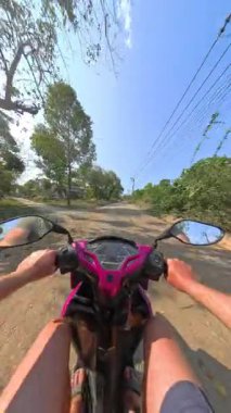 Sessiz bir kır yolu boyunca sakin bir scooter yolculuğu, her iki taraftaki yemyeşil ağaçlarla çevrili, hareketli doğanın dingin güzelliğini yakalıyor.