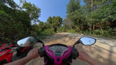 Çevresi canlı yeşil ağaçlar ve yemyeşil bitki yaşamlarıyla çevrili, orman yolunda rahat bir şekilde scooter süren bir insan, doğanın huzurunu yakalıyor.