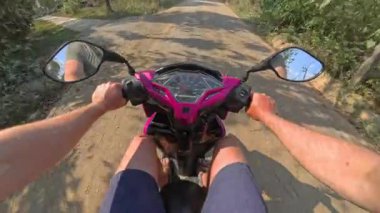Güneşli bir günde motosiklet süren bir insan, yeşilliklerle çevrili, huzurlu, asfaltsız bir kırsal yolda yol alır.