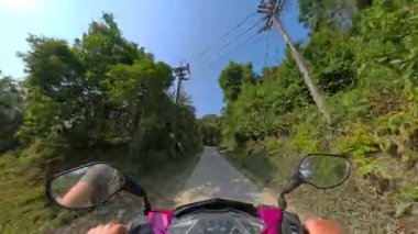 Çerçevesi yemyeşil ağaçlar ve canlı doğal çevreyle çevrili sessiz bir köy yolunda sorunsuzca giden bir scooter.