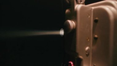 Eski bir film projektörü, karanlık odada nostaljik bir sinema parlaklığı yaratan makaralar yavaşça dönerken lensinden parlak, odaklı bir ışık huzmesi yansıtıyor.