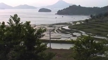 Klasik VHS görüntüleri, alacakaranlıkta sakin bir Güney Kore kıyı şeridini, sakin, ada noktalı bir denize ve arka planda sisli volkanik dağlara doğru inen teraslı pirinç tarlalarını gösteriyor.