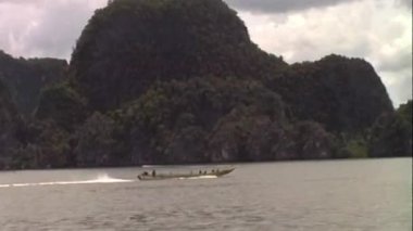 Phuket 'ten gelen bu analog görüntüler geleneksel bir uzun kuyruklu teknenin suda süzülüşünü gösteriyor, arkasında bir uyanış bırakıyor, parçalı bulutlu bir gökyüzünün altında yeşil, kayalık bir karst zeminine kurulmuş.