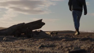 Yalnız bir adam kumlu bir sahilde, dalgalı bir dalgadan geçip ufukta siluetli bir figüre doğru yürüyor. Okyanus dalgaları yavaşça arka planda süzülüyor.