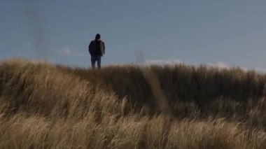 Bir adam, açık mavi gökyüzünün altında rüzgarda sallanan uzun otların arasında yavaşça yürür, sakin ve huzurlu bir doğal atmosfer yaratır.