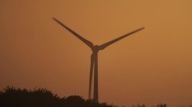 Dönen bir rüzgar türbini canlı turuncu bir günbatımı gökyüzü tarafından çerçevelendirilir, dingin doğal ortamdaki yenilenebilir enerjiyi sembolize eder.