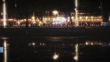 Motosikletler, canlı festival bayrakları yukarıda sallanırken ve canlı ışıklar suyu yansıtırken, canlı ama sakin bir akşam atmosferi yaratırken, sahile doğru yol alıyorlar.