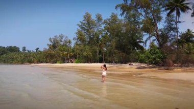 İnsansız hava aracı, el değmemiş beyaz bir tropik kumsal boyunca yürüyen genç bir kadını kristal berraklığında turkuaz dalgalarla kıyı boyunca nazikçe dolaşırken yakalar.