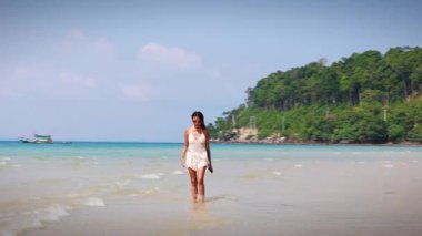 Zarif bir kavisli çekim, el değmemiş kumlu bir plajda yürüyen bir kadını, kristal berrak turkuaz okyanus ve yumuşak, güneşli dalgalar tarafından çerçevelenmiş vücudunu yakalar.