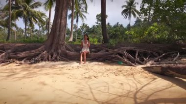 Sahildeki ağaç kökleri üzerinde oturan, telefonuna odaklanmış, yemyeşil orman yapraklarıyla çevrili bir kadının insansız hava aracı görüntüsü.