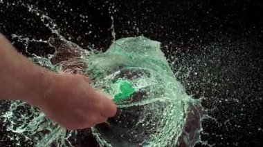 Sivri bir iğne yeşil bir su balonunu ağır çekimde patlatır. Suyu yoğun bir şekilde siyah arka plana doğru uçurur.