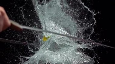 İki kılıç, sarı bir su balonunu ağır çekimde parçalayarak dramatik bir siyah arka plana doğru patlayan bir sıvıyı tetikler.