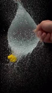 Keskin bir iğne, sarı bir su balonunu yavaş çekimde patlamanın kesin anını yakalar ve siyah arkaplana çarpar.