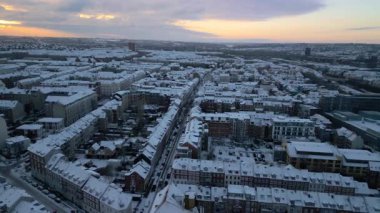 Geniş bir hava aracı görüntüsü, beyaz binalar, sokaklar ve ağaçlarla kaplanmış taze karla kaplı bir şehri yakalıyor. Yumuşak parıltının aydınlattığı sakin bir kış sahnesi yaratıyor.