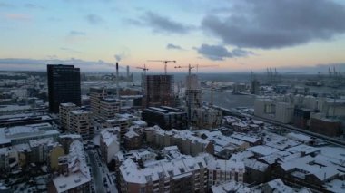 Bu süpürme hava görüntüsü, karla kaplı çatılar ve endüstriyel binaların arka planda sakin bir liman bulunan lüks gökdelenler ve vinçlerle buluştuğu canlı bir kış şehri üzerinde süzülüyor.