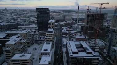 Bu insansız hava aracı görüntüsü kışın karla kaplı bir şehir merkezini yakalıyor, modern binalar ve devam eden inşaat hattı trafiğin olduğu bir ana yol, parlak, yüksek binalardan oluşan bir zemin üzerine kurulmuş.