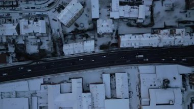 Karla kaplı çatı ve ağaçların üzerinde süzülen insansız hava aracı, hareket halindeki araçların çarpıcı bir kontrast oluşturduğu ana yolu ortaya çıkarırken, dinamik bir hava manzarası yukarıdan bir kış şehrini yakalıyor.