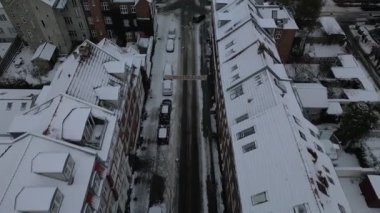 Bu İHA görüntüsü soğuk çatıları, sessiz caddeleri ve park edilmiş araçları olan soğuk bir şehir ızgarasının üzerinde süzülür. Bu arada uzak mesafede vinçleri ve gemileri olan bir endüstriyel liman vardır.