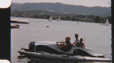 Güneşli bir günü güneşli bir gölde yakalayan nostaljik bir 8 mm film, mutlu bir ailenin pedal sürüşünün keyfini çıkarırken yelkenlilerin etraflarında yavaşça süzülmesi.