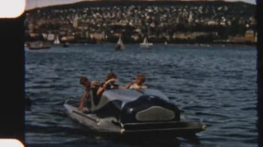 8mm 'lik klasik görüntüler, göl kenarındaki eğlenceli bir macerayı yakalıyor. Su ve uzaktaki bir şehir tarafından çerçevelenmiş, kardeşleriyle birlikte tekne turunun keyfini çıkarıyorlar.