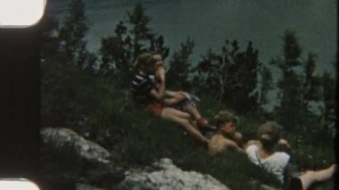 8 mm 'lik bir görüntüde çocuklar çimenli, kayalık bir tepede uzanmış, göl manzaralı, doğa gezisi sırasında mola veriyorlar.