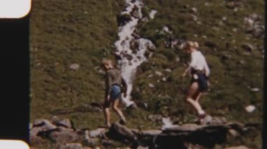 Güneşli bir günde etrafı çimlerle çevrili sığ, taşlarla dolu bir nehri dikkatle geçen bir ailenin 8 mm 'lik görüntüsü.