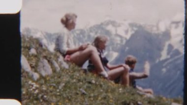 Uzakta karla kaplı tepeleri olan çimenli bir vadide dinlenen çocukların 8mm 'lik klasik klibi.