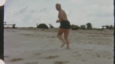 1970 'lerin Tunus' undan 8mm 'lik bir film, güneşin kumlu kıyısı boyunca koşan bir adamı gösteriyor. Soldan gelen hafif dalgalar ve seyrek palmiye ağaçları ve plaja gidenler gibi ayak izleri bırakıyor.