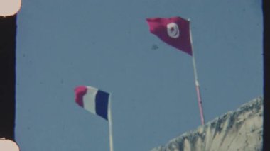 1970 'lerin Tunus' undan çekilen bu 8 mm 'lik film, soluk mavi gökyüzüne karşı yıpranmış bir ışık taşı yapının üzerinde Fransız ve Tunus bayraklarının düşük açılı çekimini gösteriyor.