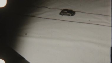 Klasik 8mm stop motion klipte koyu renk paralel çizgilerle işaretlenmiş hafif kumaş üzerinde hareket eden küçük siyah oyuncak araba görünür çıkıntı delikleriyle güçlendirilmiş kumlu, eski ve eğlenceli atmosfer yaratır.