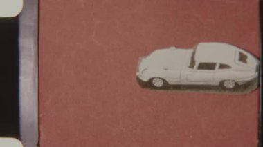8mm stop motion animasyon çerçevesinde kırmızı-kahverengi desenli arka planda hareket eden beyaz bir klasik arabanın stilize edilmiş kesimini gösteriyor.