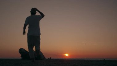 A romantic couple stands on a rock by the ocean, watching the sunset over the horizon
