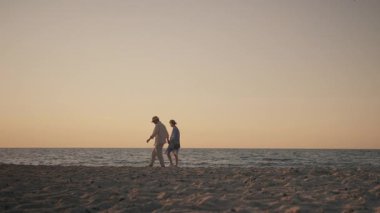 Static wide shot of a romantic couple walking hand in hand along a sandy beach at sunset