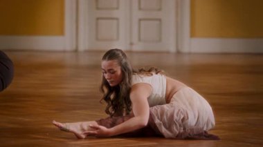 A young female dancer gracefully stretches her leg in a beautifully lit hall with elegant parquet flooring, showcasing her flexibility and focus