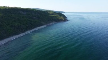 A wild empty sandy beach between a rocky sharp large ledge, covered with green fresh new vegetation, and a deep calm cool Black Sea under a clear blue sky. UHD 4K video realtime