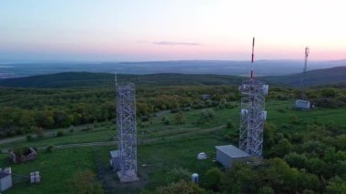Birds eye view of hills covered with green spring vegetation with tall professional cell towers, against the backdrop of farmhouse blooming planted fields and a clear blue sky. UHD 4K video realtime