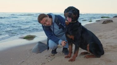 A caring adult fair-haired girl in a warm gray tracksuit combs her beloved big guard dog of the Rottweiler breed, on a sandy wild empty beach near the calm blue Black Sea